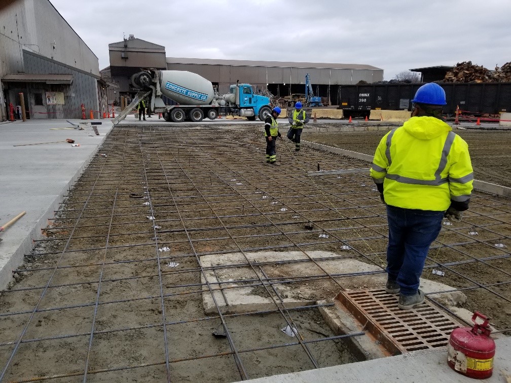 A concrete truck is seen in the background, and multiple workers are doing measurements to prepare to fill a large spot with concrete paving.
