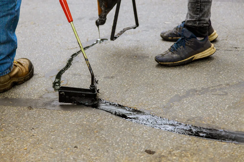 The shoes of two workers can been seen standing on either side of a crack in the asphalt pavement. They are filling the crack to fix it.